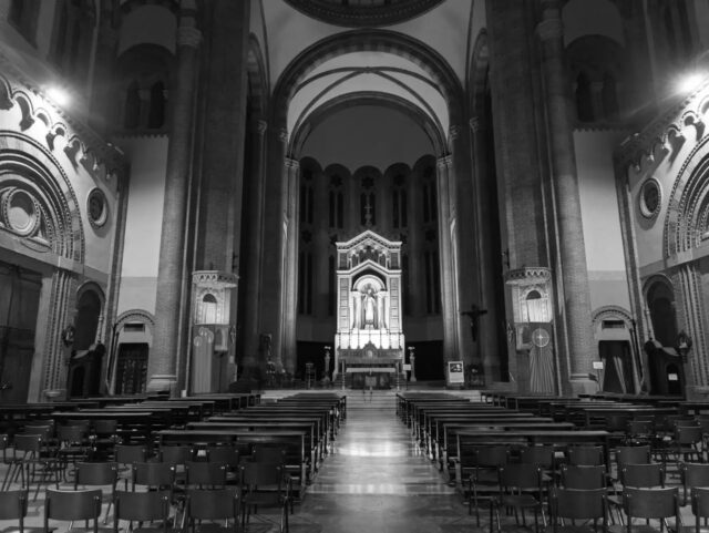 Cartoline dalla Bolognina – 2026
Dentro il Sacro Cuore

Dall'esterno è una mole che si vede dai treni. Dentro, la sorpresa è che non c'è nessuna delle cose che ci si aspetta da una chiesa grande: niente navate laterali, niente colonne, niente file di pilastri che accompagnano lo sguardo verso l'altare. C'è un'unica aula. Cinquantadue metri dalla porta al fondo dell'abside, ventinove di larghezza media, e sopra la testa la croce della cupola a sessanta metri.

È una scelta precisa. Edoardo Collamarini disegna il progetto nel 1897 su una pianta centrale, con riferimenti dichiarati a Santa Sofia di Costantinopoli e allo stile neobizantino del Sacré-Cœur di Montmartre: siamo nel pieno dell'eclettismo di fine Ottocento, quando le chiese nuove si costruiscono citando l'Oriente cristiano invece del gotico. L'assemblea non è incolonnata, è raccolta sotto un vuoto.

I lavori partono nel 1901 e vanno per gradi: il tempio apre al culto nel 1912, e le decorazioni interne arrivano solo fra il 1913 e il 1914. Da lì si legge la stratigrafia. Il ciborio è disegnato dallo stesso Collamarini, gli affreschi dell'abside sono di Domenico Ferri, il trittico scultoreo dell'altare maggiore è di Arturo Orsoni. Il trittico dipinto di Leopoldo Perucchi porta la data del 1915, l'anno in cui la chiesa diventa parrocchia autonoma per servire un rione che nel frattempo si era riempito.

Poi il crollo della cupola nel 1929, il rifacimento concluso nel 1935, i danni del bombardamento del 1943 e la ricostruzione fino al 1947: quello che si guarda oggi ha quattro età diverse. Le uniche cose davvero antiche sono due opere lignee ai lati del presbiterio, un bassorilievo con la Madonna e un Crocifisso databili fra Cinque e Seicento, arrivate qui da altrove.

La prima parte a essere completata fu la cripta, nel 1903. Le prime case popolari della Bolognina sono del 1908: qui la chiesa è arrivata prima delle case.