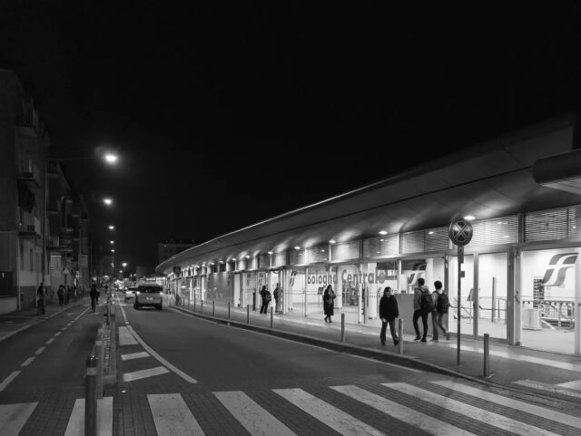 Cartoline dalla Bolognina – 2026
Via de' Carracci

Per un secolo e mezzo questa non è stata una strada, ma un retro. Quando la stazione apre nel 1864, l'attuale via de' Carracci segna già il limite nord del piazzale ferroviario: da una parte i binari, i capannoni di servizio, il deposito locomotive; dall'altra, ancora campagna. Il fronte era di là, verso piazza delle Medaglie d'Oro. 

Di qua c'era il muro.

Il piano del 1889 costruisce la Bolognina addossata proprio a quel muro, perché è la ferrovia a dare lavoro. E per la stessa ragione il rione paga: Bologna è un nodo ferroviario, e nella Seconda guerra mondiale subisce trentadue bombardamenti pesanti. La Bolognina viene colpita duramente il 24 agosto 1944.

Nel dopoguerra il lato Carracci resta quello che era: mercato ortofrutticolo, scalo merci, capannoni. Poi chiudono anche quelli, e per vent'anni il rione si ritrova davanti un grande vuoto urbano, su cui si progetta molto — c'è pure un piano di Ricardo Bofill — senza costruire niente.

Il cambio arriva con l'Alta Velocità. Fra il 2004 e il 2007, prima di costruire, si scava per capire cosa c'è: emergono una necropoli romana, una strada glareata, canalizzazioni e fornaci. Poi, dove correvano i binari 12, 13, 14 e 15, viene scavata a cielo aperto una stazione profonda ventitré metri. L'8 giugno 2013 viene inaugurata.

Chi ci abita ricorda soprattutto il rumore, le vibrazioni e la polvere di anni di cantiere. E non è finita: nel luglio 2026 sono partiti i lavori per completare l'asse Nord-Sud, atteso da vent'anni.

La strada che per centocinquant'anni ha tenuto la Bolognina dietro la ferrovia adesso ci passa sotto.