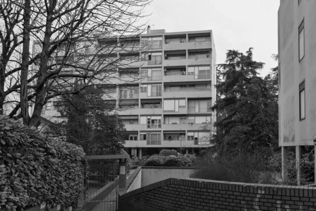 Cartoline dalla Bolognina – 2026
La casa in Bolognina

Se si guarda la Bolognina dall'alto si capisce subito una cosa: qui vive un pezzo di città molto più grande di quanto lo spazio lasci immaginare. 

Sono 36.681 persone, più della metà dell'intero quartiere Navile e quasi un decimo di tutta Bologna. La densità lo dice meglio di qualunque descrizione: fra i 97 e i 120 abitanti per ettaro, contro una media cittadina di 28. Nei punti più compressi la Bolognina è affollata quanto il centro storico.

Ci si è arrivati in due tempi. Prima gli isolati chiusi del piano del 1889, quattro piani più il solaio, costruiti sul filo della strada con il cortile dentro. Poi, dal dopoguerra, il modello si rovescia: l'edificio si stacca dalla strada, si pianta in mezzo al lotto e sale fino a otto piani. Dove stavano otto famiglie ne stanno molte di più.

Il risultato sono più di ventimila abitazioni, terza zona di Bologna per numero dietro San Vitale e Mazzini. E sono le più piccole della città: 82 metri quadri di media, contro i 108 di Santo Stefano e i quasi 150 dei Colli.

Piccole, però, non vuol più dire economiche. A luglio 2026 nella zona Bolognina-Arcoveggio-Navile si chiedevano in media 3.522 euro al metro quadro, il nove per cento in più rispetto all'anno prima. La media cittadina sta sopra i 3.800: il rione non è più il posto dove si risparmia, è appena sotto la media.

Del resto lo spazio libero, qui, non c'è mai stato. Il Comune scrive che è difficile arrivare a una dotazione adeguata di verde e di scuole. E i redditi mediani più bassi di tutta Bologna si registrano in due sole zone: questa e San Donato.

Le case più piccole della città, nel rione dove si guadagna meno, oggi costano il nove per cento in più di un anno fa