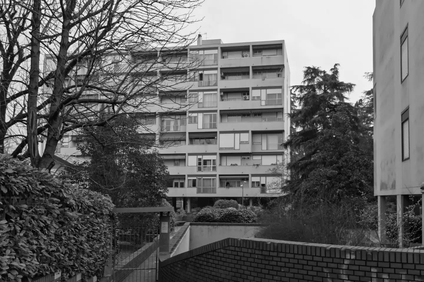 Cartoline dalla Bolognina – 2026
La casa in Bolognina

Se si guarda la Bolognina dall'alto si capisce subito una cosa: qui vive un pezzo di città molto più grande di quanto lo spazio lasci immaginare. 

Sono 36.681 persone, più della metà dell'intero quartiere Navile e quasi un decimo di tutta Bologna. La densità lo dice meglio di qualunque descrizione: fra i 97 e i 120 abitanti per ettaro, contro una media cittadina di 28. Nei punti più compressi la Bolognina è affollata quanto il centro storico.

Ci si è arrivati in due tempi. Prima gli isolati chiusi del piano del 1889, quattro piani più il solaio, costruiti sul filo della strada con il cortile dentro. Poi, dal dopoguerra, il modello si rovescia: l'edificio si stacca dalla strada, si pianta in mezzo al lotto e sale fino a otto piani. Dove stavano otto famiglie ne stanno molte di più.

Il risultato sono più di ventimila abitazioni, terza zona di Bologna per numero dietro San Vitale e Mazzini. E sono le più piccole della città: 82 metri quadri di media, contro i 108 di Santo Stefano e i quasi 150 dei Colli.

Piccole, però, non vuol più dire economiche. A luglio 2026 nella zona Bolognina-Arcoveggio-Navile si chiedevano in media 3.522 euro al metro quadro, il nove per cento in più rispetto all'anno prima. La media cittadina sta sopra i 3.800: il rione non è più il posto dove si risparmia, è appena sotto la media.

Del resto lo spazio libero, qui, non c'è mai stato. Il Comune scrive che è difficile arrivare a una dotazione adeguata di verde e di scuole. E i redditi mediani più bassi di tutta Bologna si registrano in due sole zone: questa e San Donato.

Le case più piccole della città, nel rione dove si guadagna meno, oggi costano il nove per cento in più di un anno fa