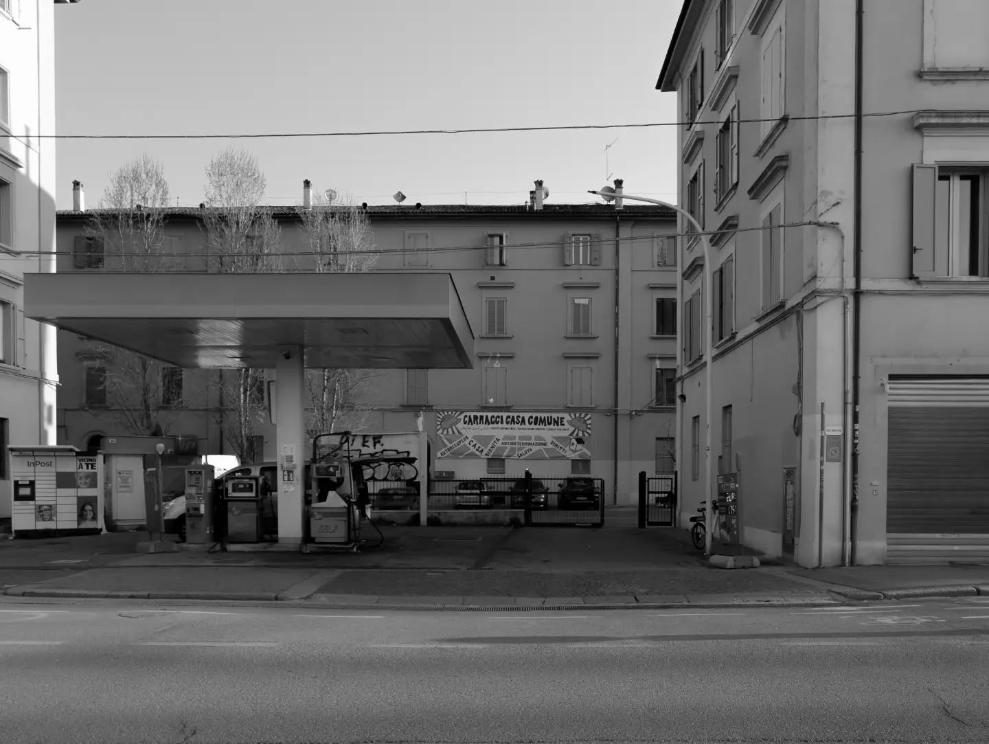 Cartoline dalla Bolognina – 2026
Carracci Casa Comune

Nell'ottobre del 2023 tre palazzine pubbliche vuote in via de' Carracci 63, a due passi dai binari, vengono occupate. Ci entrano oltre centodieci persone — età diverse, provenienze diverse, molti bambini — che il mercato della casa a Bologna aveva semplicemente espulso.

L'occupazione prende un nome: Carracci Casa Comune.
Non è un gesto improvvisato. Il percorso è accompagnato da Plat, la Piattaforma di intervento sociale che a Bologna si occupa di diritto all'abitare, e si intreccia con l'area di Laboratorio Bologna: assemblee, presidi, presenza costante in città, una trattativa tenuta aperta con l'amministrazione mese dopo mese. E dentro le palazzine si organizza una comunità che si regge da sé, decide in assemblea e gestisce lo stabile come una cosa collettiva. Da qui il nome: non "occupazione", casa comune.

Nella seconda metà di quell'anno a Bologna ci sono altre occupazioni abitative, in via Raimondi, in via di Corticella, in via San Giacomo. Finiscono tutte con uno sgombero. Qui no.

Sulle palazzine pende un sequestro e lo sgombero è annunciato, ma nel luglio 2024 arriva invece una convenzione fra Comune e Acer: gli edifici non vengono venduti, ma ristrutturati a spese pubbliche; ventiquattro appartamenti vanno alla transizione abitativa e poi assegnati per trent'anni con un bando; le famiglie entrano nelle graduatorie per la casa popolare. E intanto restano dove sono.

A Bologna un'occupazione che finisce con un riconoscimento e non con la polizia non succedeva dal 1983.

Dietro la stazione, sulle scale, restano i pezzi delle reti di ferro che sbarravano il passaggio. Non servono più: le porte le hanno aperte dall'interno.